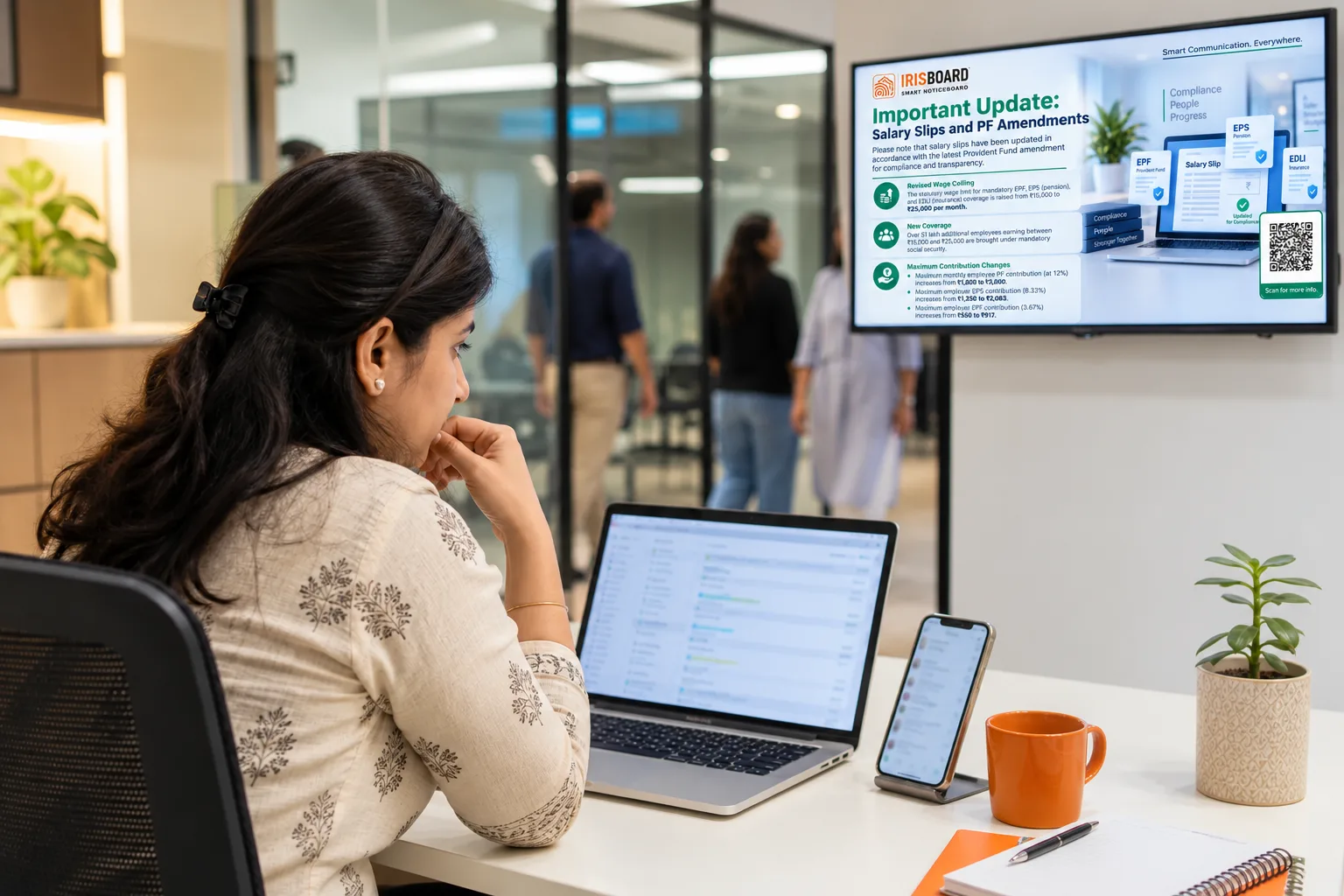 An HR executive at her desk looking at an email inbox and a phone, with a wall-mounted screen glowing in the corridor behind her